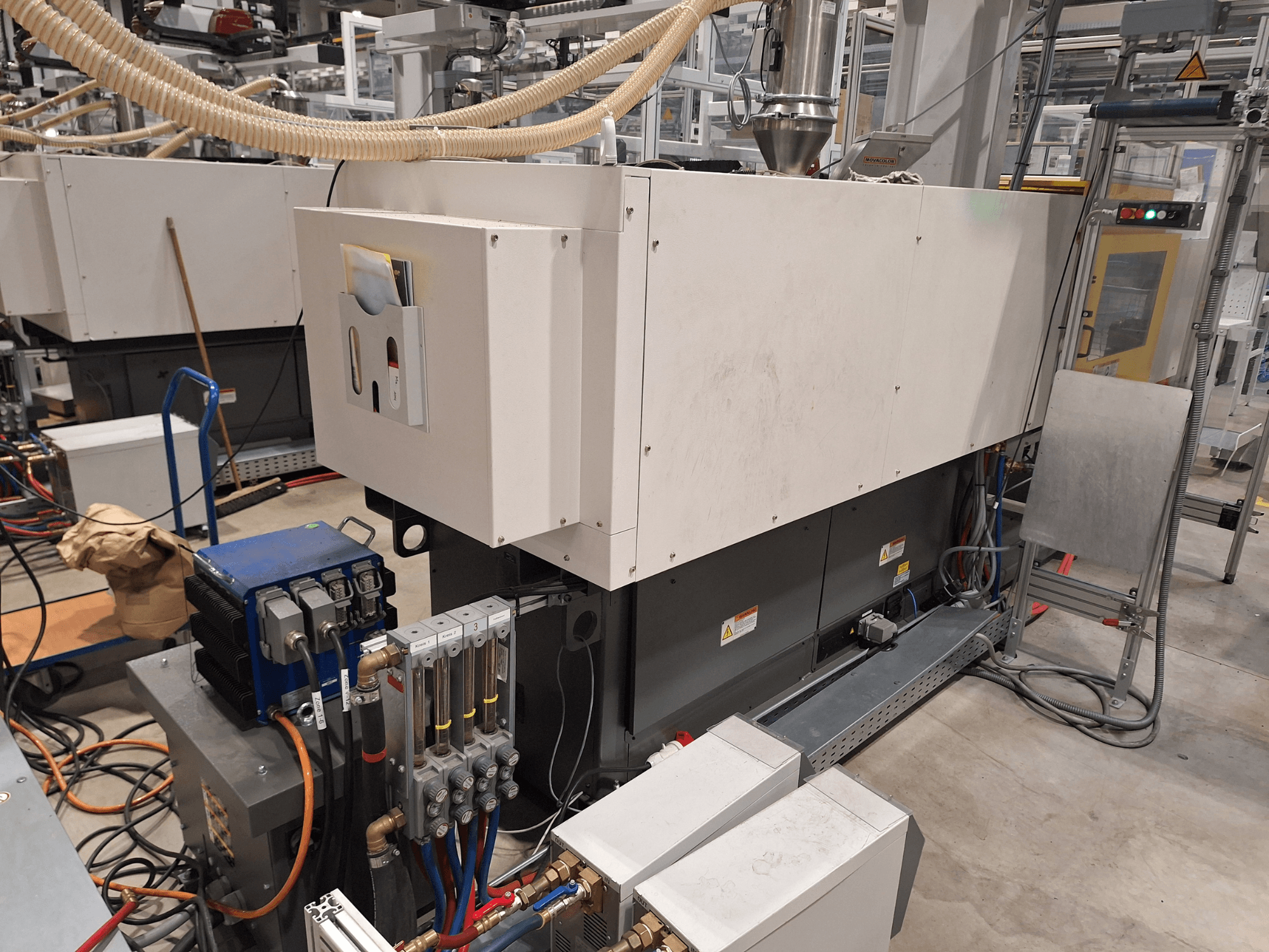 Průmyslové zařízení v továrním prostředí; zahrnuje velký stroj s ovládacím panelem, hadicemi a elektrickými přípojkami, pohled z boku.