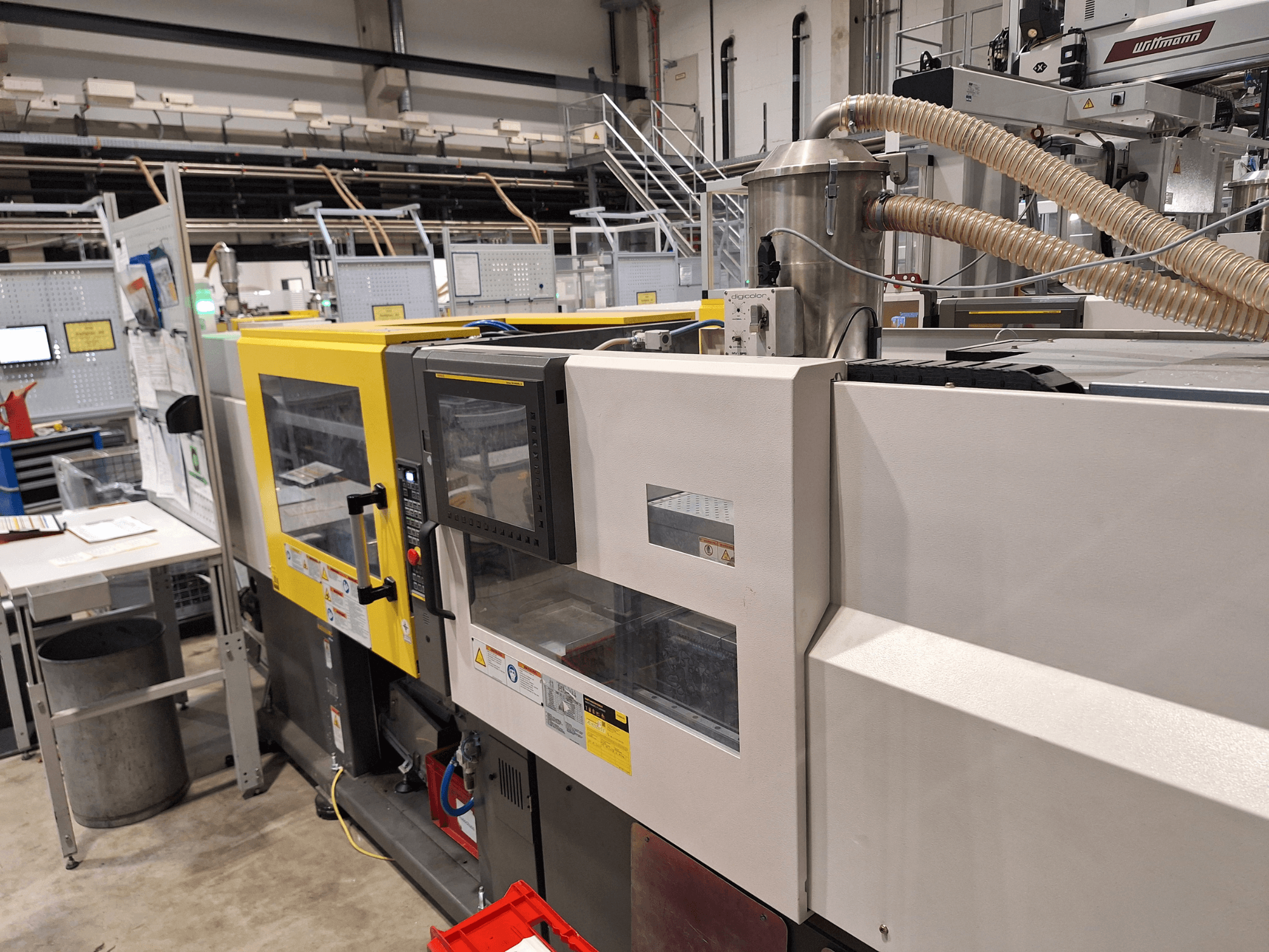 Pohled zepředu na vícebarevný vstřikovací stroj se žlutými a šedými částmi, s ovládacím panelem a výfukovým systémem.