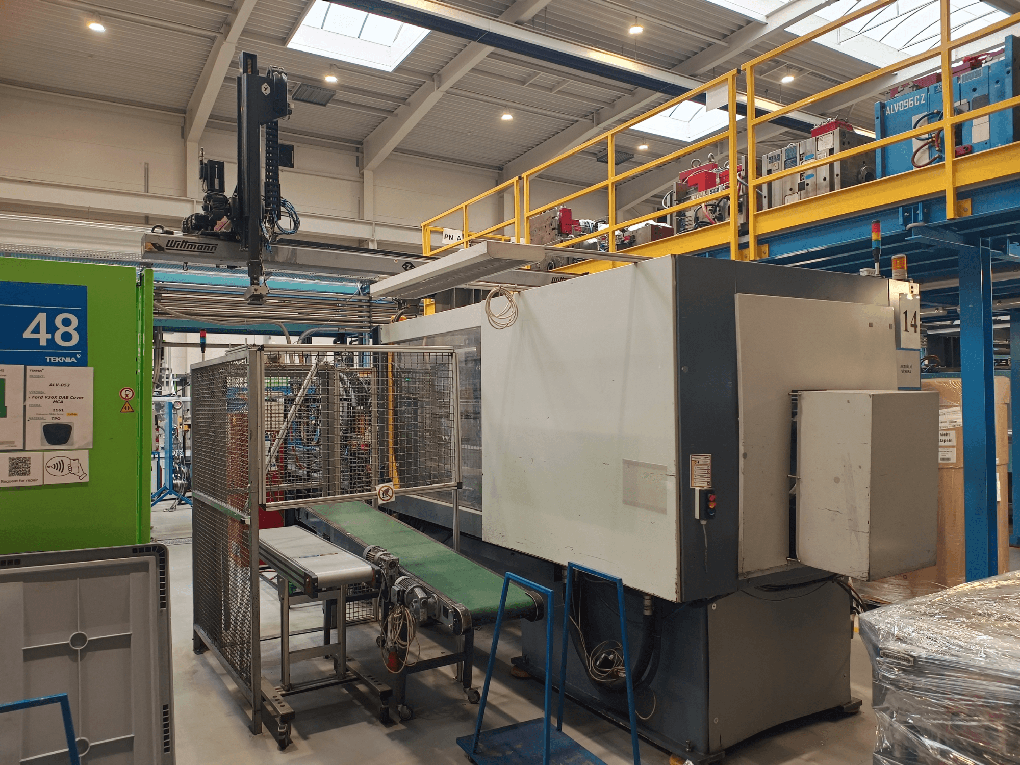 Průmyslové automatizační zařízení se zeleným dopravníkem s označením "48" a mostovým jeřábem; boční pohled na stroj v tovární hale.